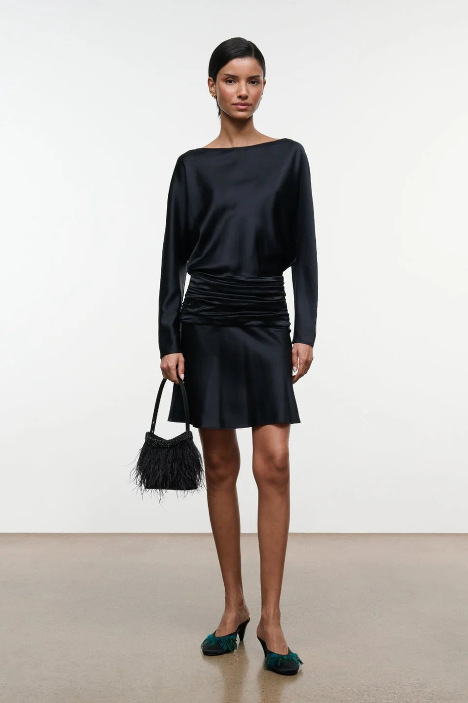 A model stands against a plain light gray studio background wearing a black long-sleeve mini dress with a draped waist, holding a small black feather-trim handbag and wearing dark green pointed-toe heels.