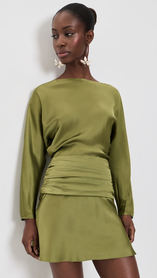 A woman modeling a sage green long-sleeve mini dress with a gathered ruched waist, photographed against a plain light gray studio background.