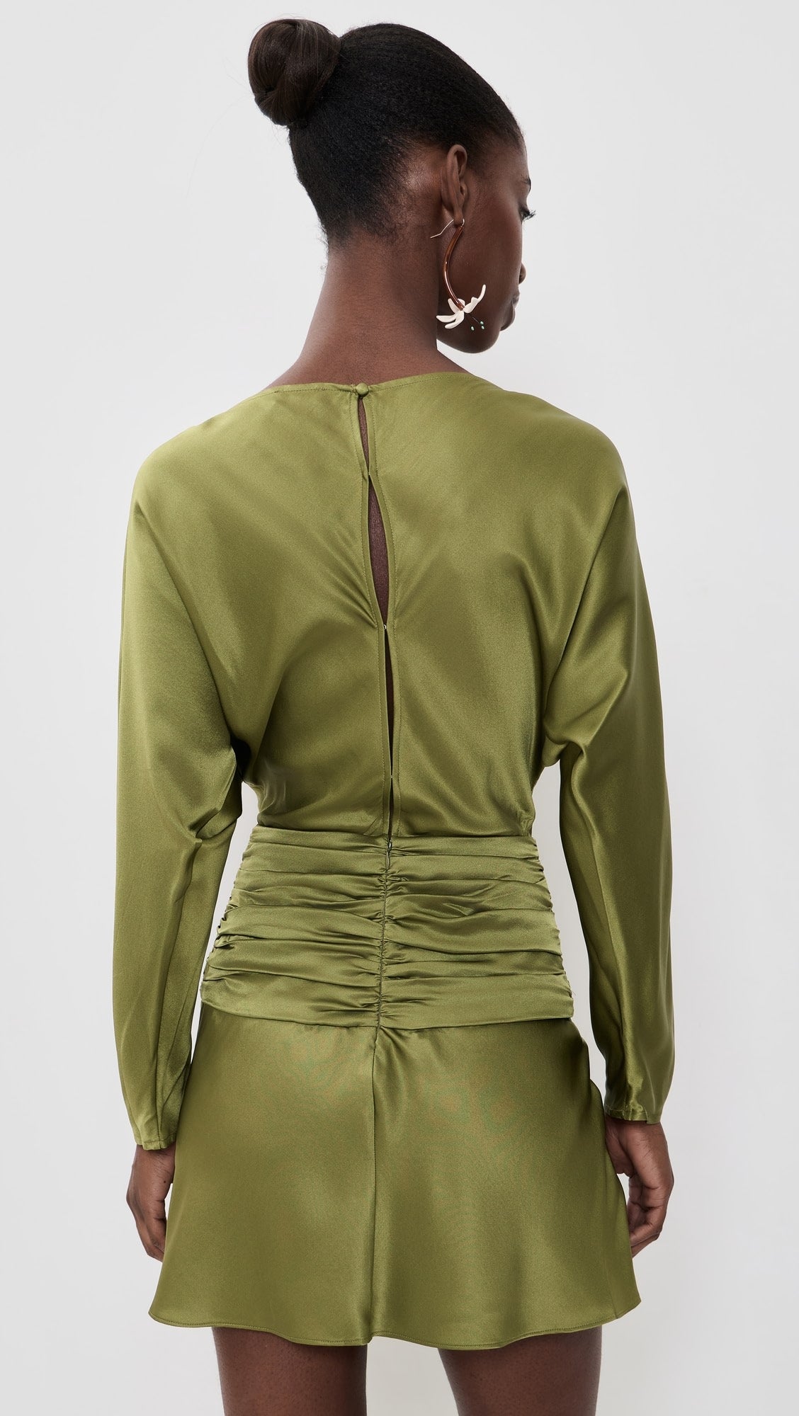 A woman shown from the back wearing a sage green long-sleeve mini dress with ruching at the waist and a small keyhole cutout at the back, photographed against a plain light gray studio background.