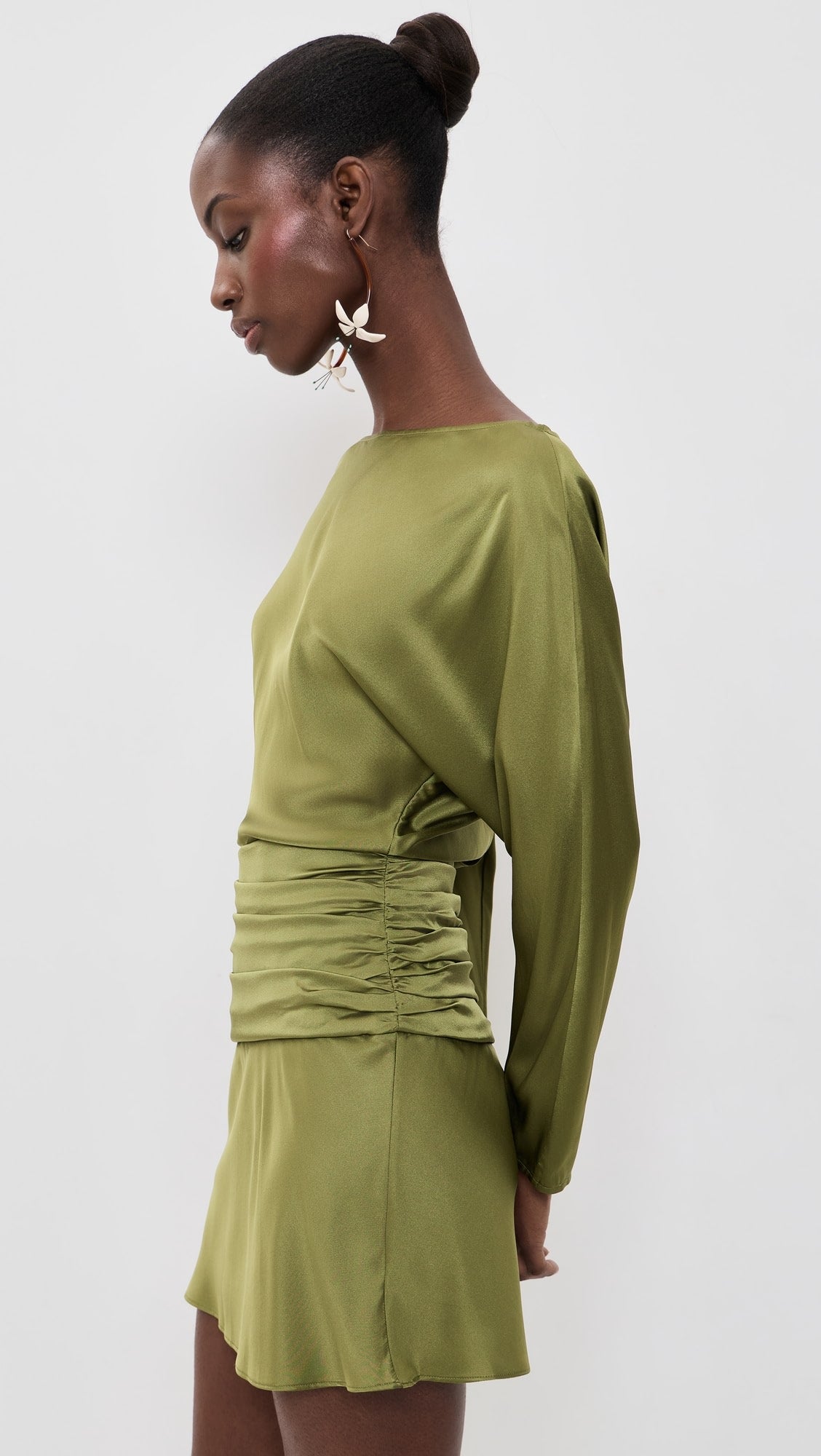 A woman modeling a sage green long-sleeve mini dress with a gathered ruched waist, photographed against a plain light gray studio background.