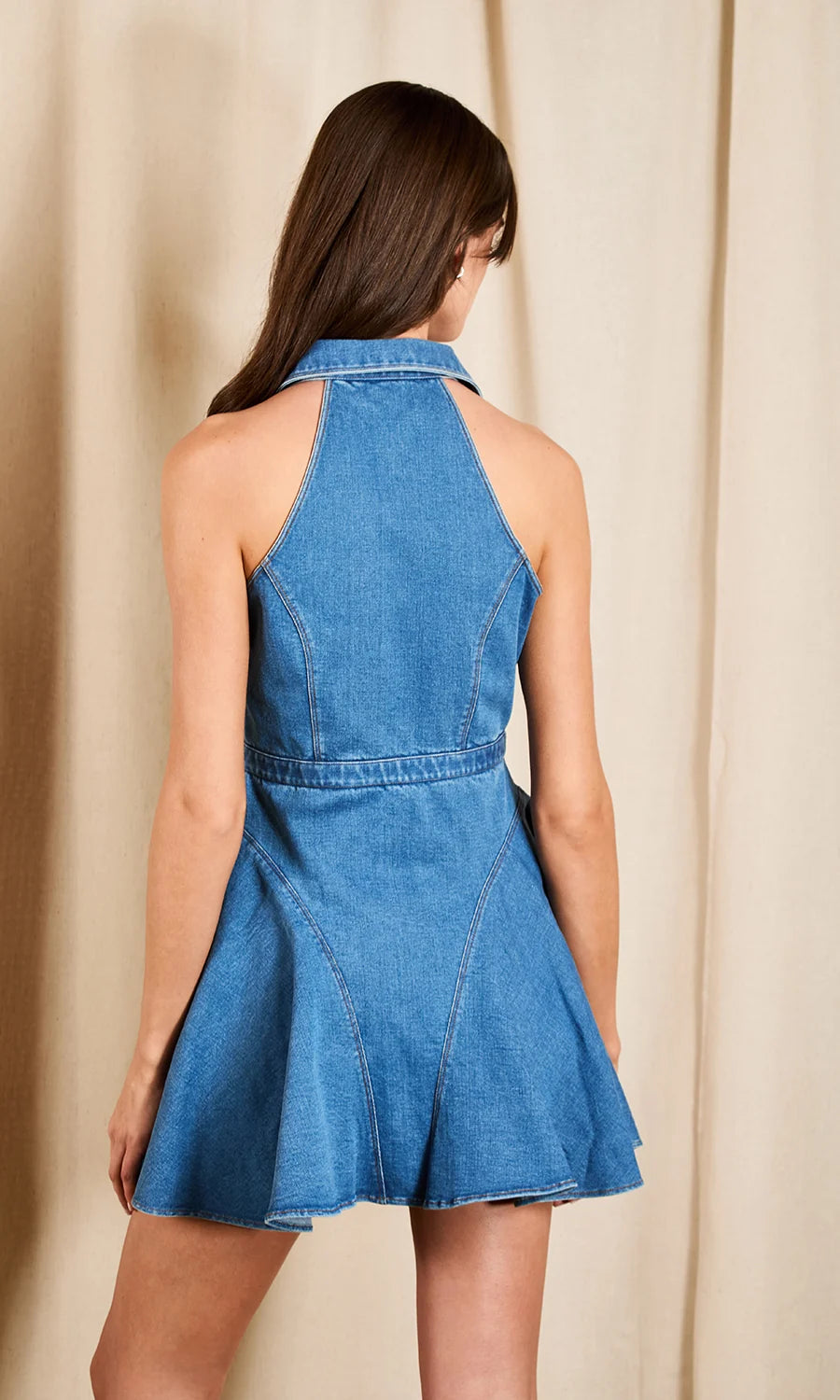 A woman wearing a blue denim halter mini dress, shown from the back against a neutral beige curtain backdrop.
