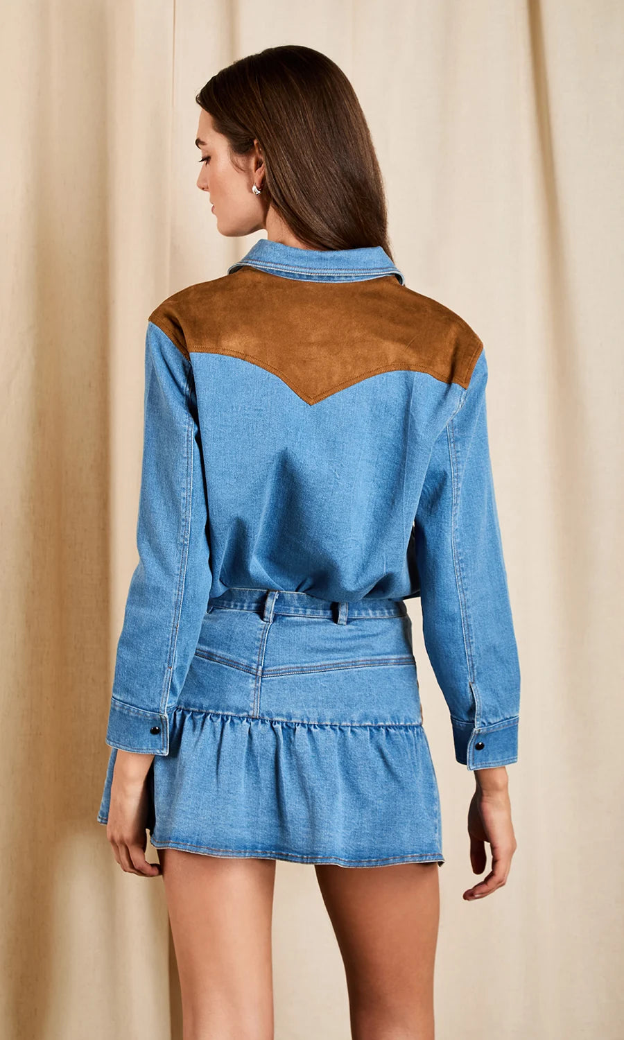 A woman shown from the back wearing a blue denim long-sleeve shirt dress with brown suede-like shoulder panels, photographed against a beige curtain backdrop.
