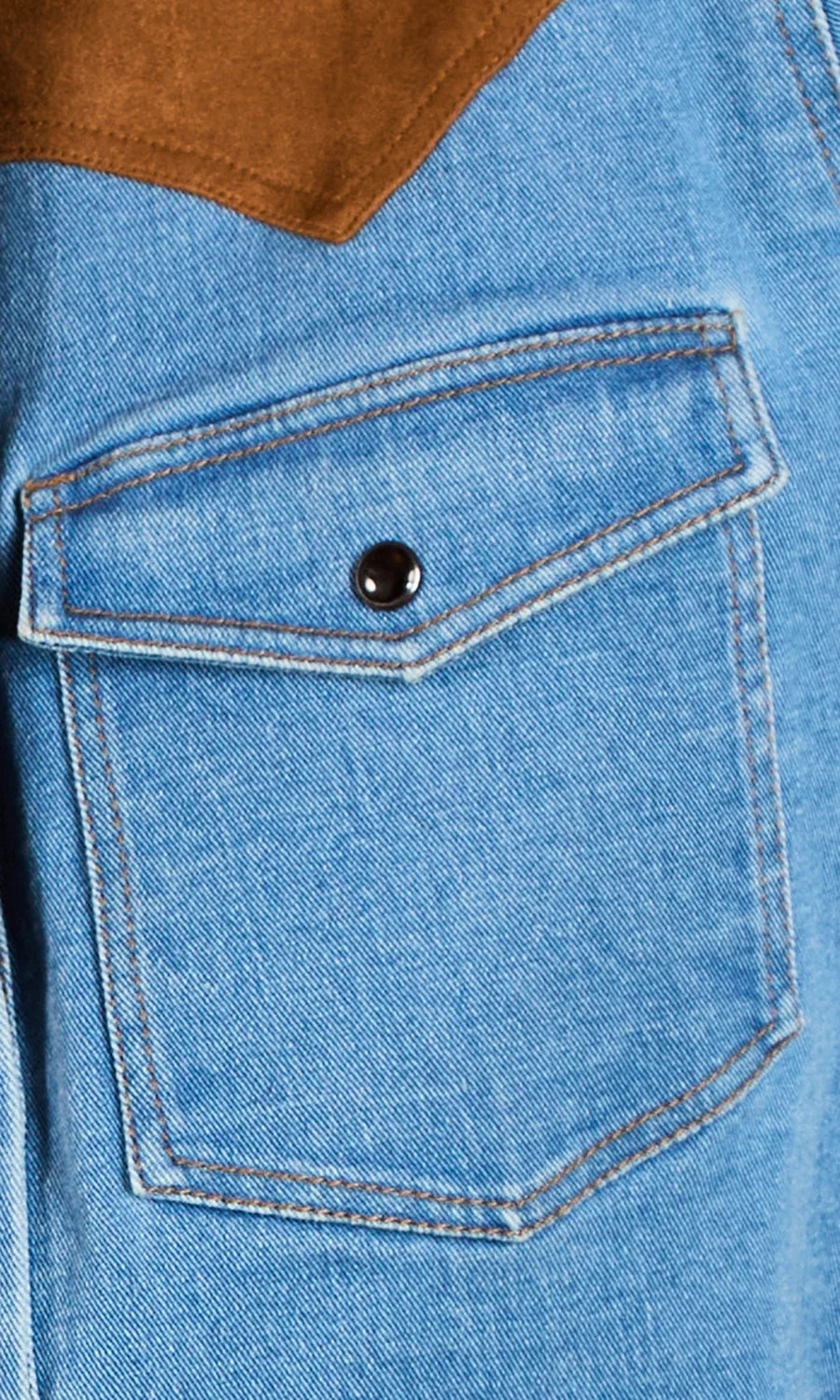 Close-up of a light blue denim jacket featuring a front flap pocket with a black snap button and a brown suede-like shoulder panel.