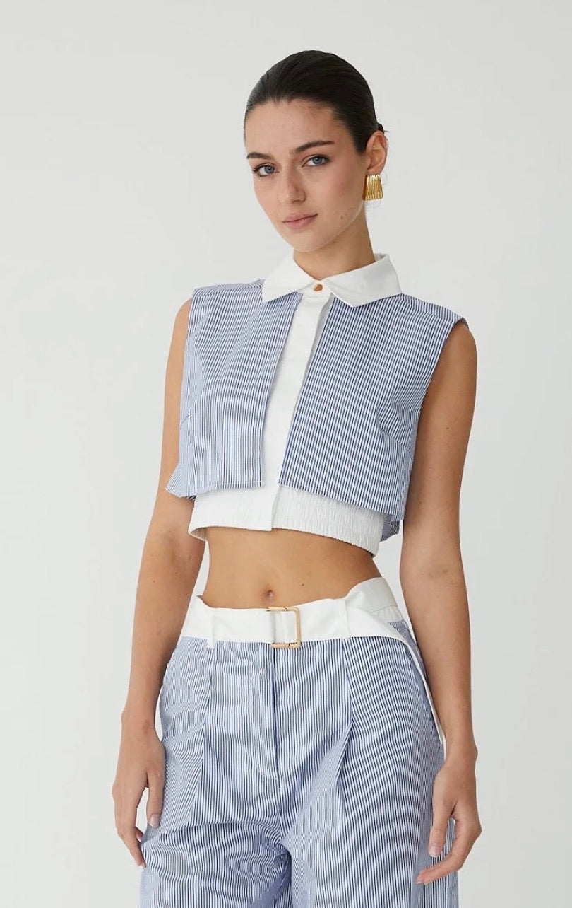 A model wearing a light blue and white striped sleeveless vest with a white collar, paired with matching striped trousers and a white belt, against a light gray studio background.