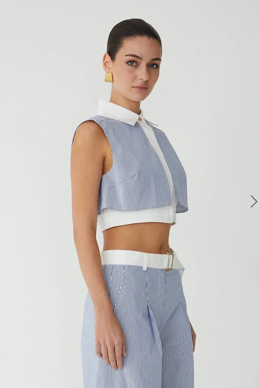 A model wearing a light blue and white striped sleeveless crop top with a white collar and white trim, paired with matching striped trousers and a white belt, against a plain light gray background.