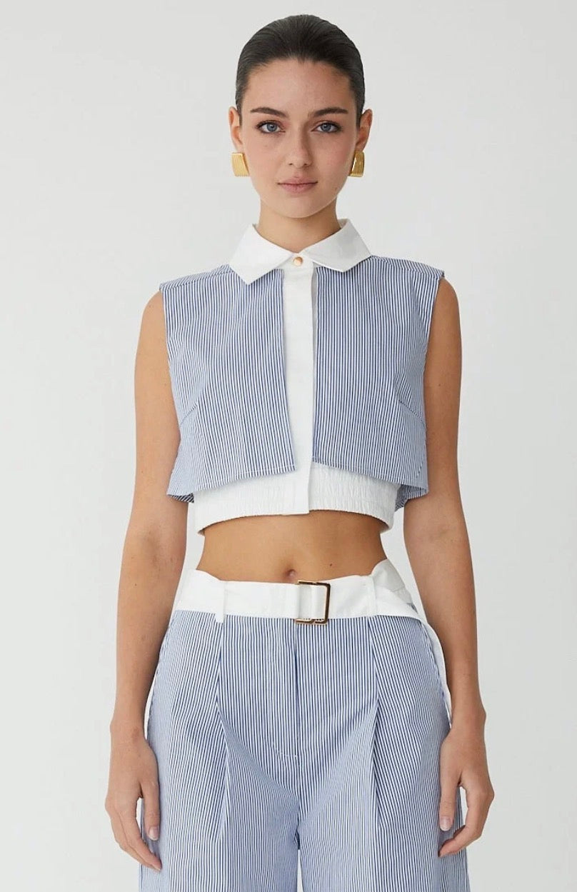 A model wearing a sleeveless light blue and white striped crop top with a white collar and white placket, paired with matching striped trousers and a white belt, against a light gray studio background.
