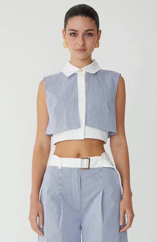 A model wearing a sleeveless light blue and white striped crop top with a white collar and white placket, paired with matching striped trousers and a white belt, against a light gray studio background.