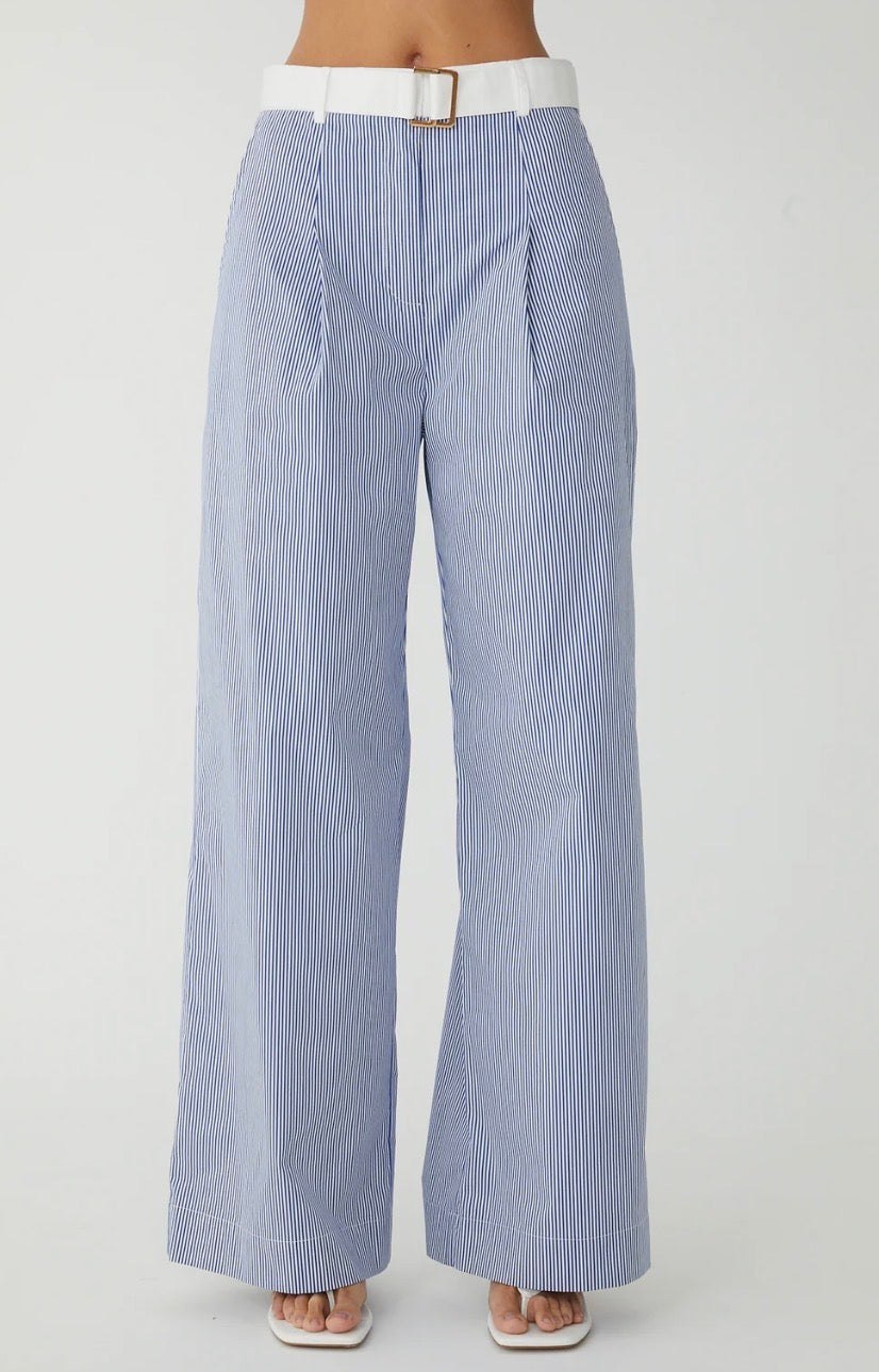 A person wearing light blue and white vertically striped wide-leg trousers with a white belt and white sandals, shown from the waist down against a plain light gray studio background.