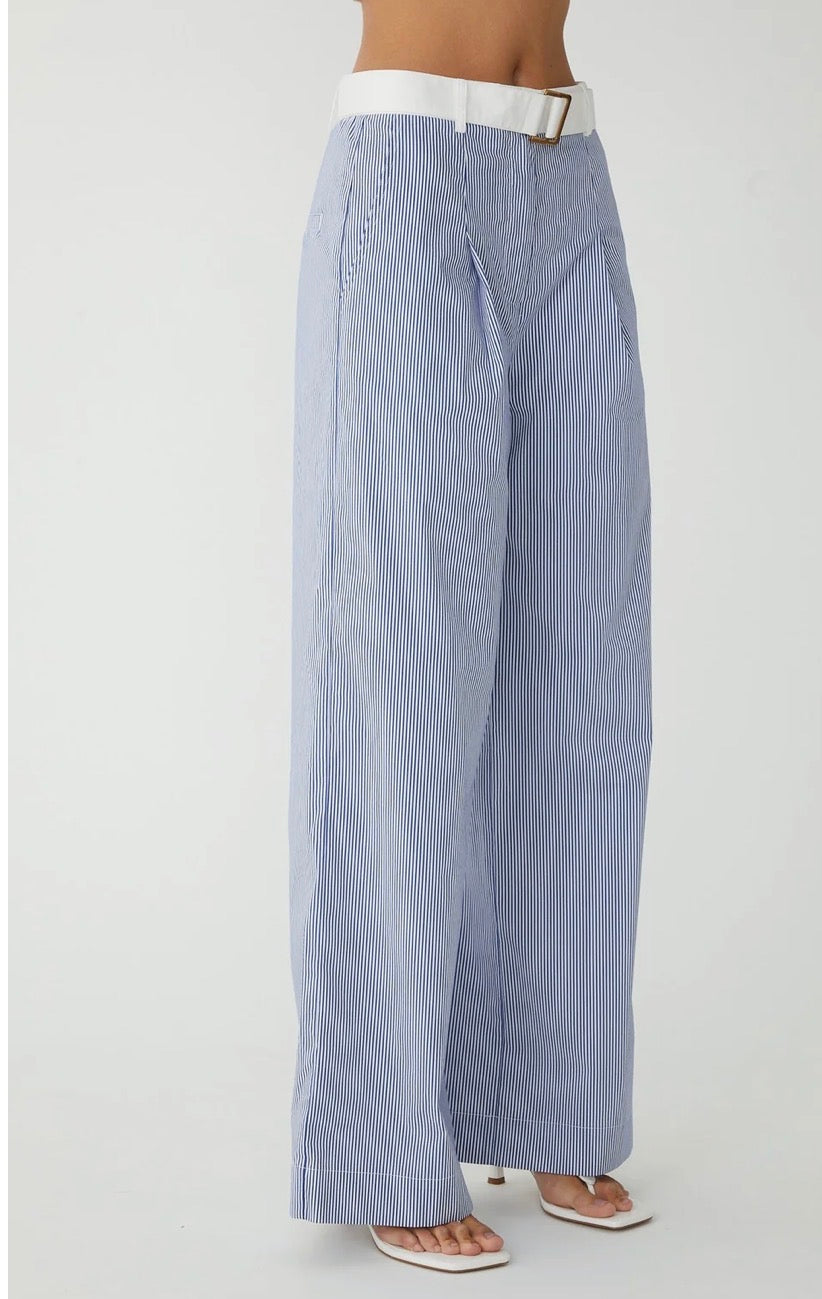 A person wearing light blue and white vertically striped wide-leg trousers with a white belt and white heeled sandals, shown from the waist down against a plain light gray studio background.