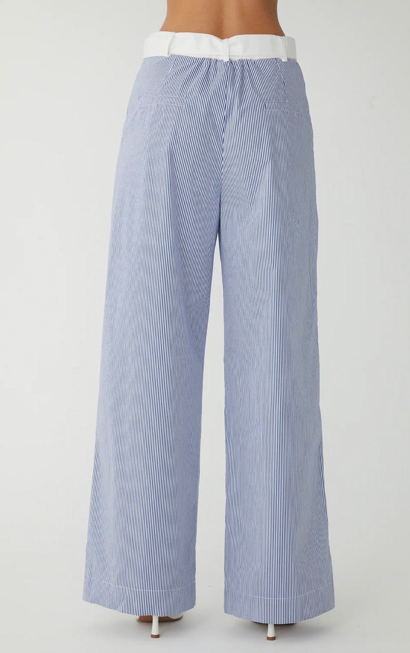 A pair of wide-leg trousers in a light blue and white vertical stripe pattern, shown from the back on a model. The trousers feature a contrasting white waistband with a small gold-tone ring detail.
