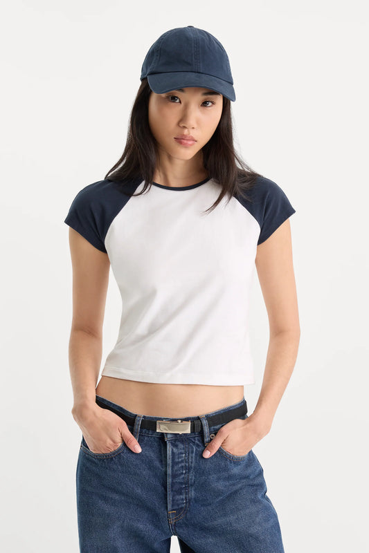 A woman modeling a white crop top with dark navy blue raglan sleeves and a matching navy baseball cap, paired with blue jeans against a plain white studio background.