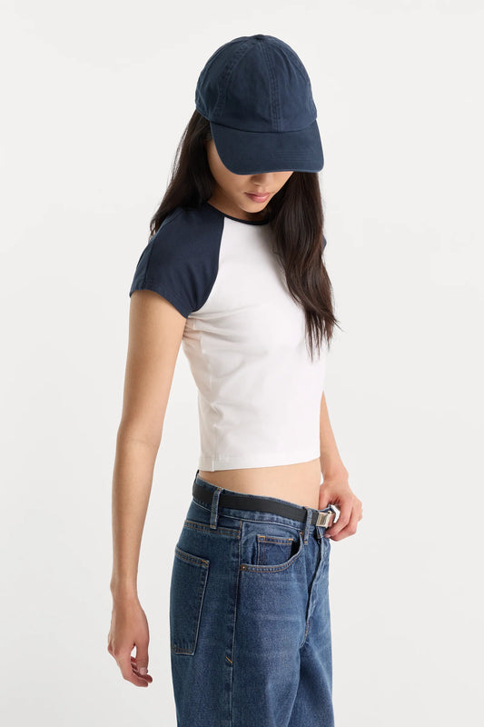 A model wearing a white crop top with navy blue raglan sleeves, a navy baseball cap, and blue jeans, photographed against a plain light gray studio background.