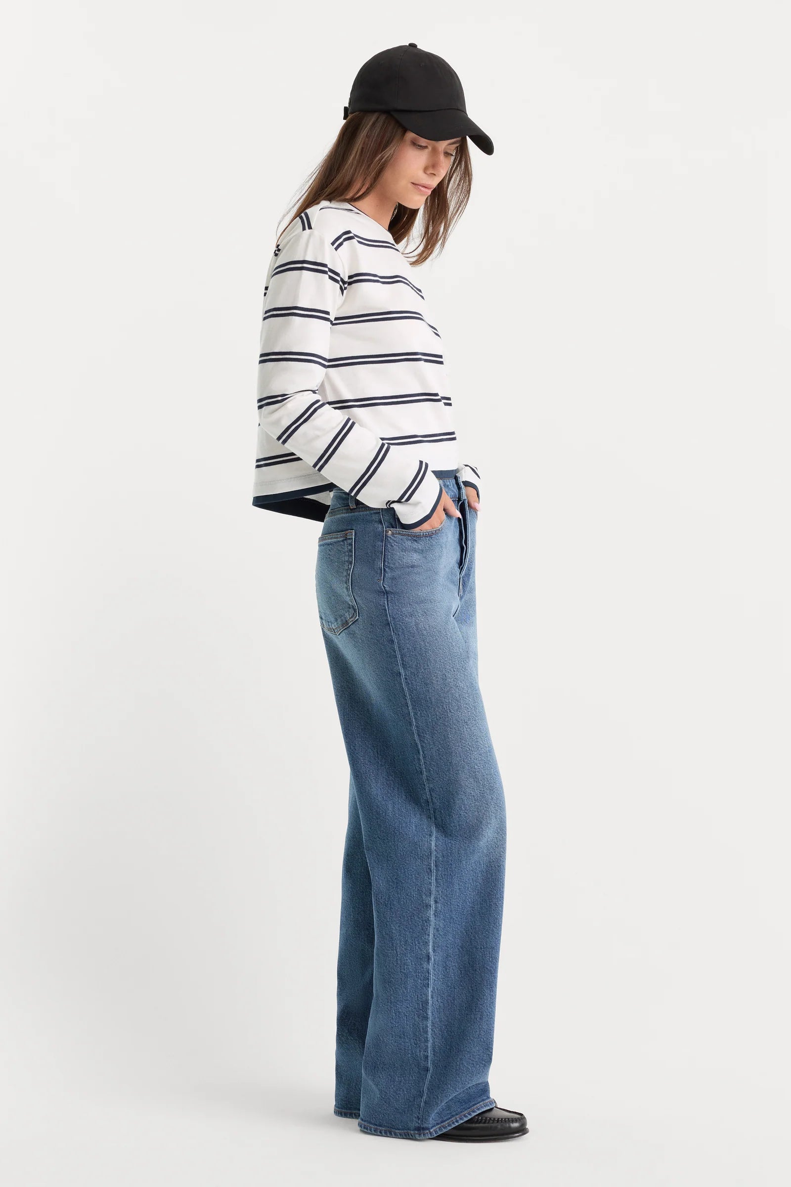 A model wearing a white long-sleeve top with thin navy horizontal stripes, high-waisted wide-leg blue jeans, and a black baseball cap, standing against a plain white studio background.