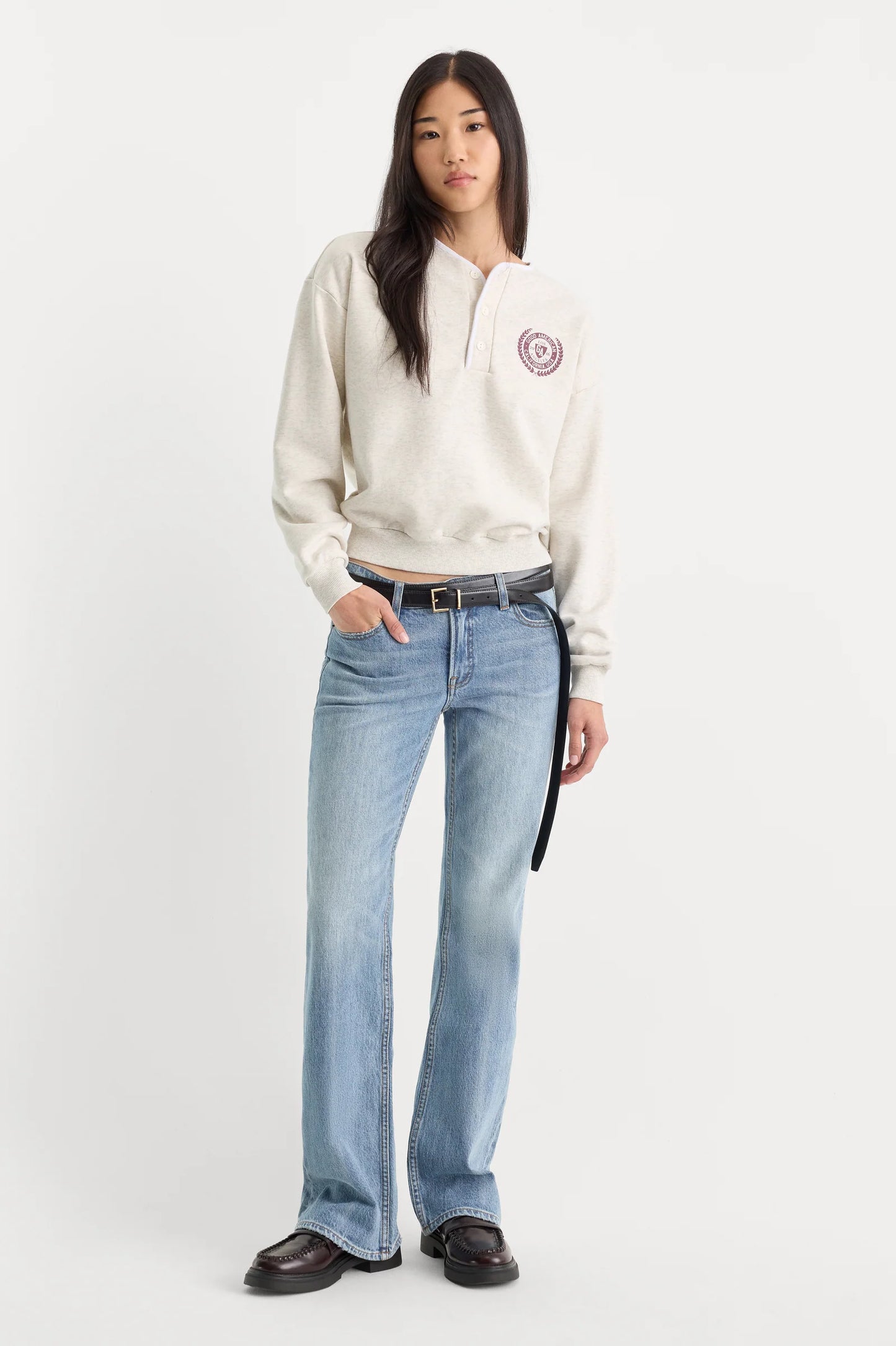A model stands against a plain light gray studio background wearing a cream-colored quarter-zip sweatshirt with a circular logo on the left chest, light-wash blue jeans, a black belt, and black loafers.
