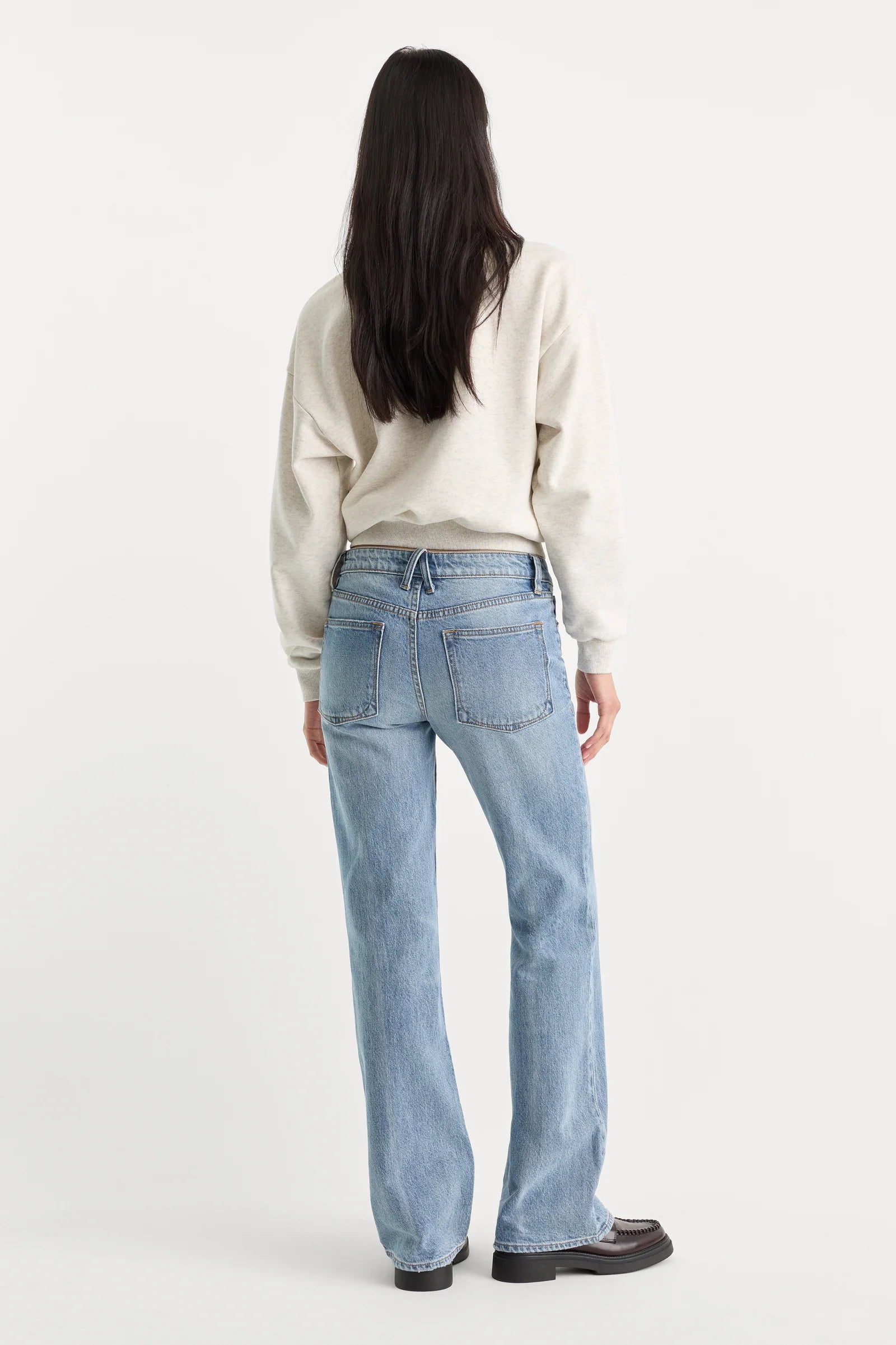 A woman viewed from behind wearing light-wash blue straight-leg jeans, a cream sweatshirt, and dark brown shoes against a plain white studio background.