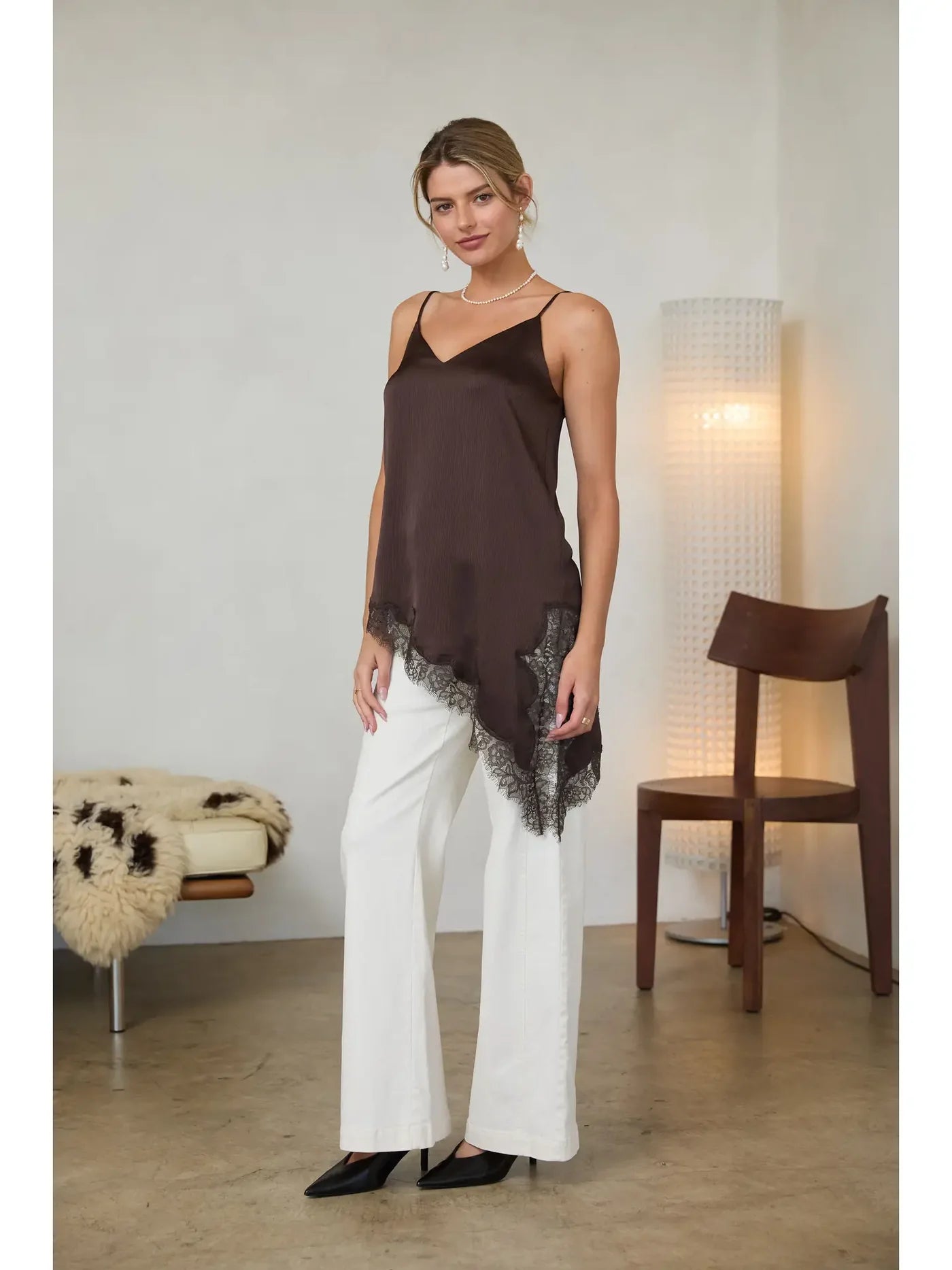 A woman models a dark brown camisole top with a black lace hem, paired with white wide-leg pants and black heels, standing in a minimalist room with a floor lamp and wooden chair.