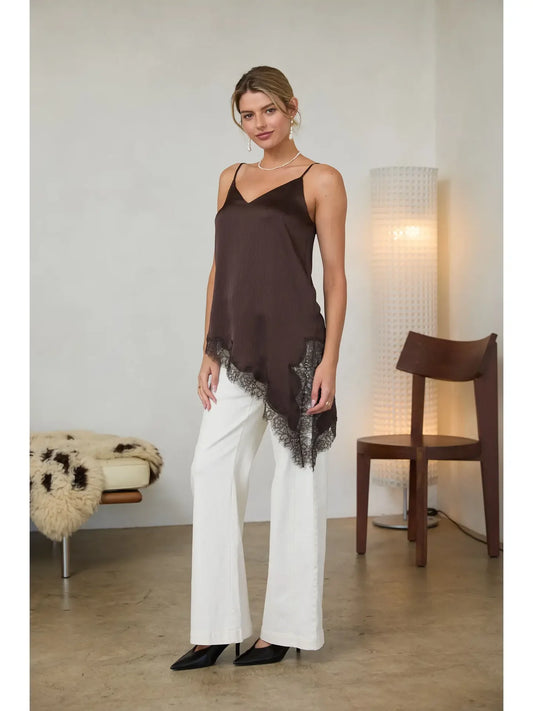 A woman models a dark brown camisole top with a black lace hem, paired with white wide-leg pants and black heels, standing in a minimalist room with a floor lamp and wooden chair.