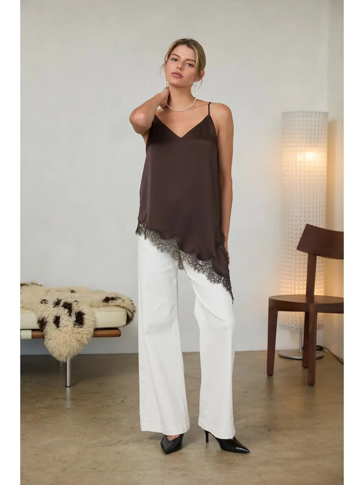 A woman models a dark brown camisole top with a sheer black lace hem, paired with white wide-leg pants and black heels, standing in a modern minimalist room.
