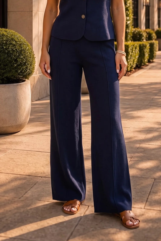 A person wearing navy blue wide-leg pants and a matching sleeveless top, standing outdoors on a sunlit stone walkway with trimmed hedges and a planter in the background.