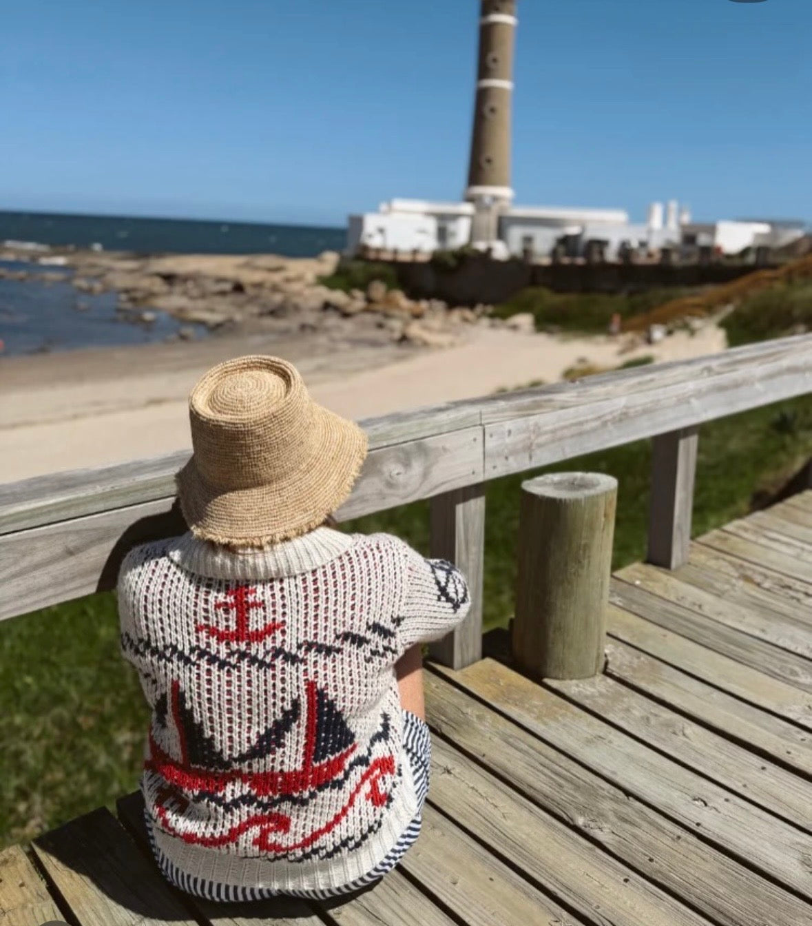A child wearing a straw sun hat and a cream knit sweater with a red and black nautical pattern sits on a wooden boardwalk overlooking a beach and lighthouse.