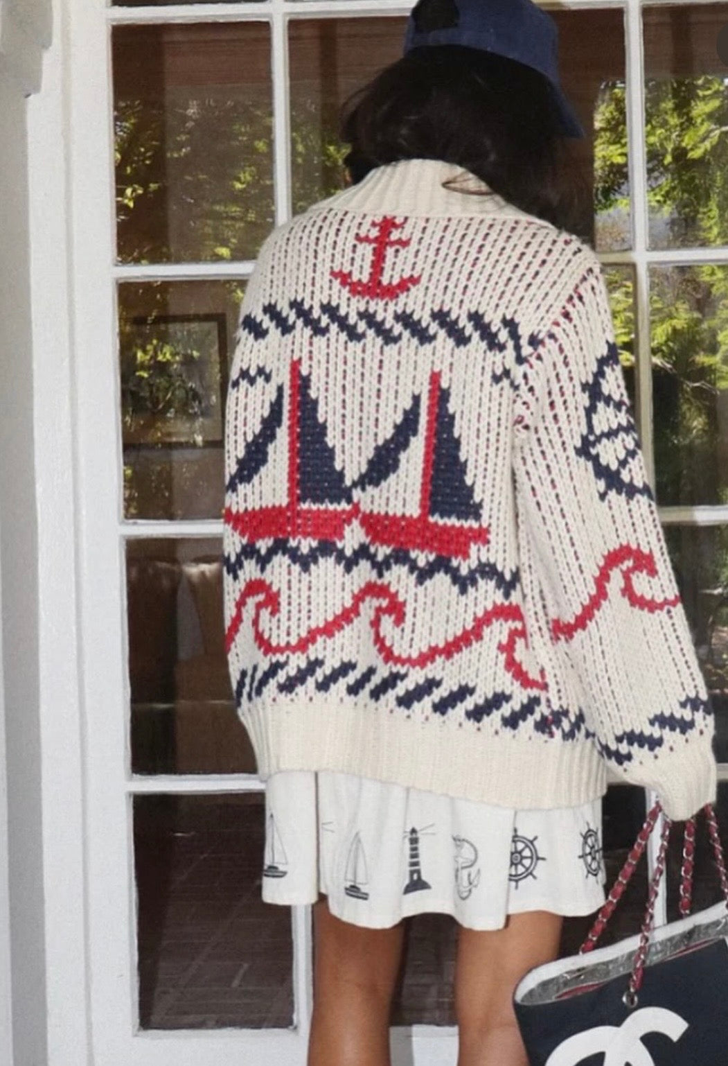 A person viewed from behind wearing a cream knit cardigan with a nautical pattern featuring red and navy sailboats, waves, and an anchor, paired with a white skirt and a navy cap.