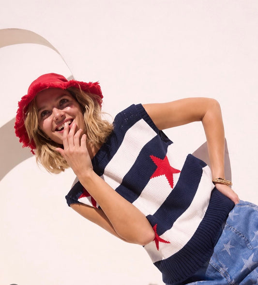 A woman wearing a red frayed bucket hat, a navy and white striped knit sweater with red stars, and blue jeans with white star prints, posed against a light beige background with a curved white architectural element.