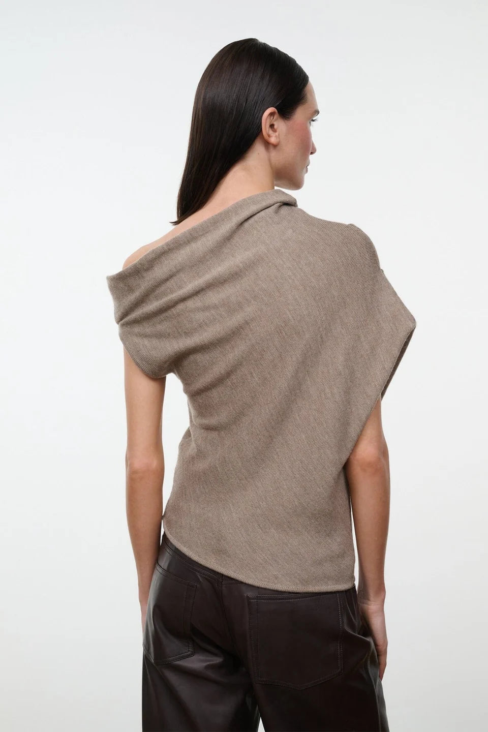 The back view of a model wearing the STAUD Mabel one shoulder sweater in sesame with brown leather pants on a neutral background.