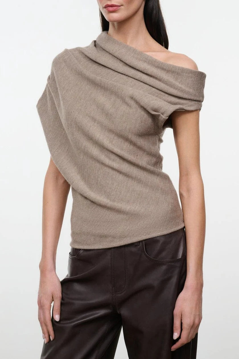 Closeup of a model wearing the STAUD Mabel one shoulder sweater in sesame with brown leather pants on a neutral background.