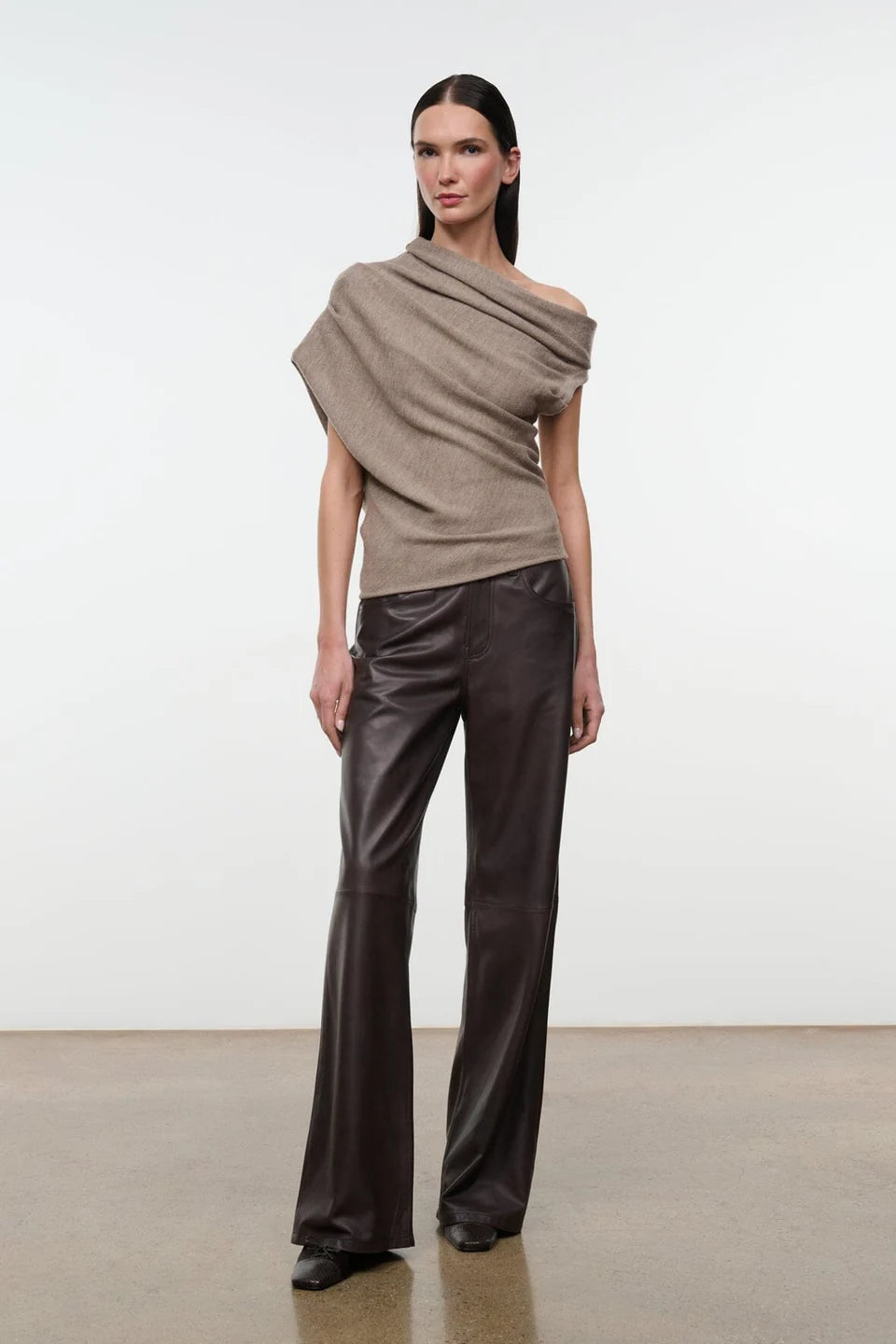 Model wearing the STAUD Mabel one shoulder sweater in sesame with brown leather pants on a neutral backkground.