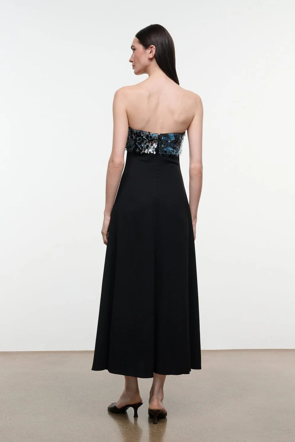 Model from the back view wearing the STAUD Phoenix ankle length black dress that features a straight strapless neckline with an embellished shiny top showing the zipper closure on a neutral background.