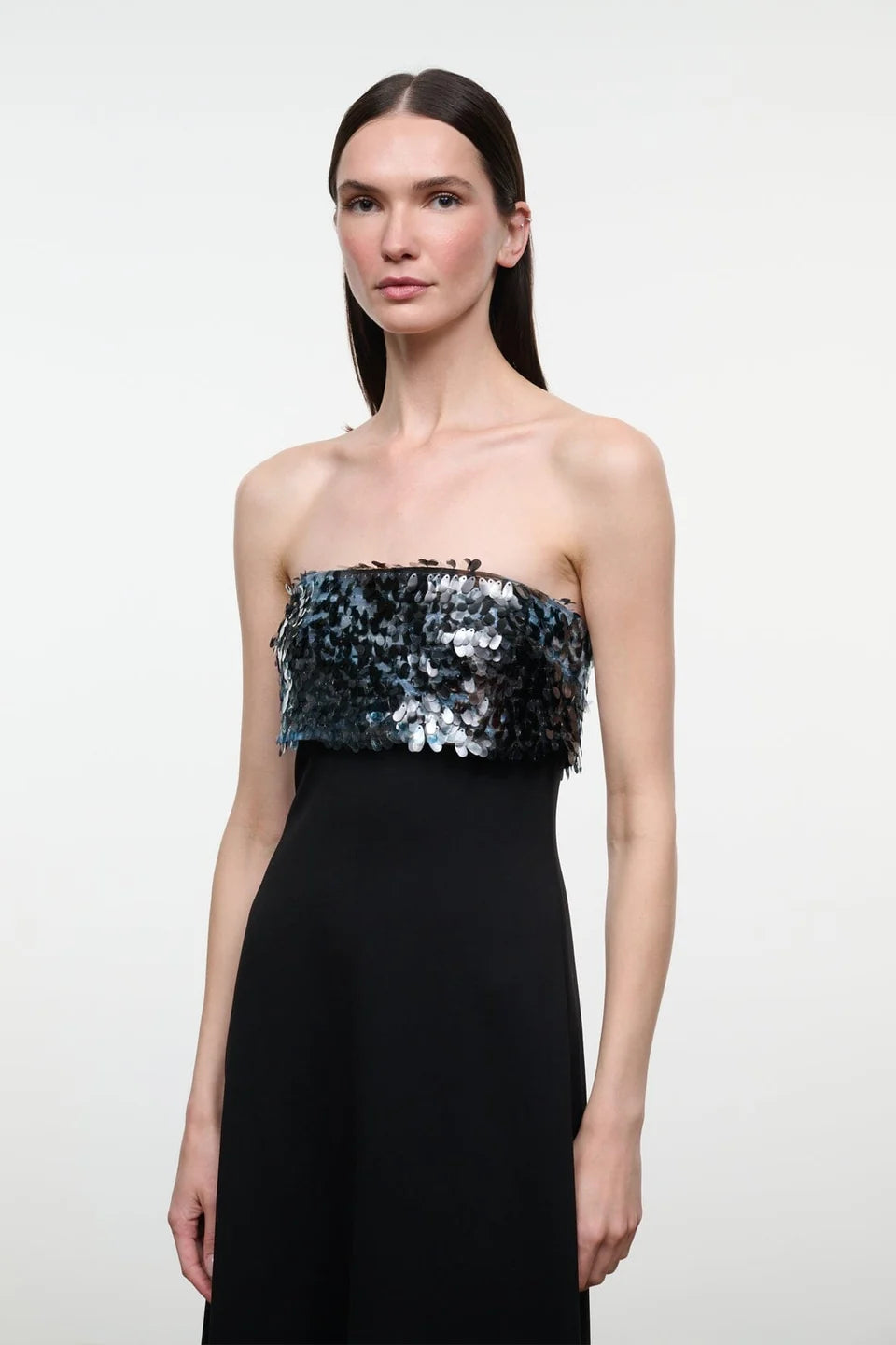 A closeup of a model wearing the STAUD Phoenix ankle length black dress that features a straight strapless neckline with an embellished shiny top on a neutral background.