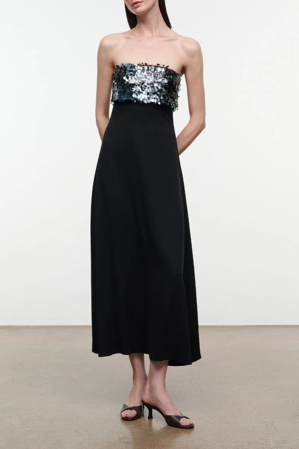 Model wearing the STAUD Phoenix ankle length black dress that features a straight strapless neckline with an embellished shiny top on a neutral background.