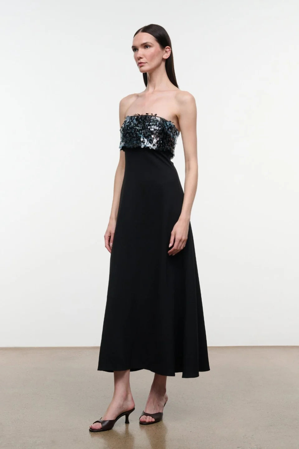 Model wearing the STAUD Phoenix ankle length black dress that features a straight strapless neckline with an embellished shiny top on a neutral background.