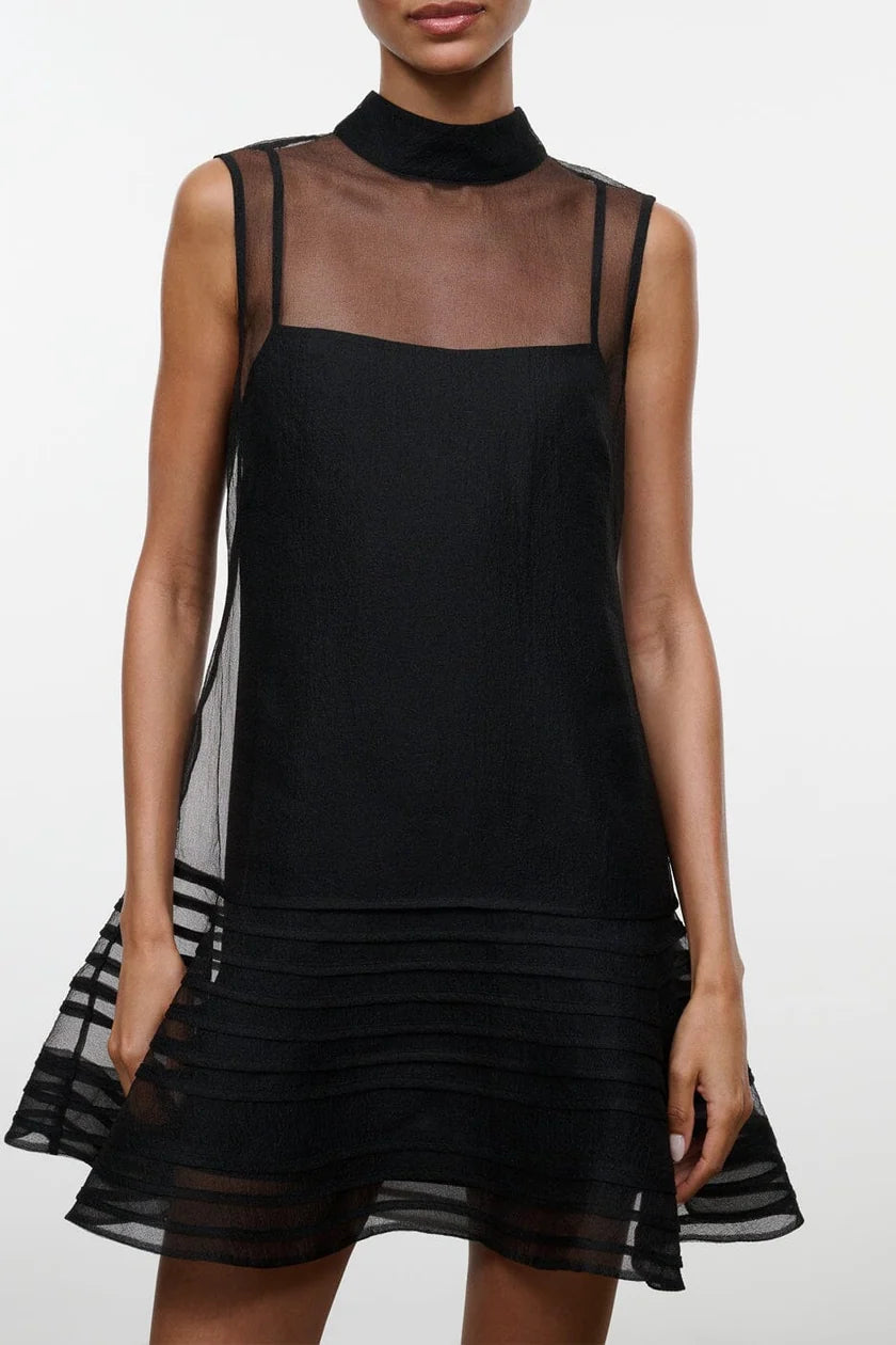 Model on a neutral background wearing the STAUD sterling mini dress in black which features a sheer outer layer with a high neck and a mini black slip underneath. The closeup displays the horizontal line detail on the bottom of the outer layer.