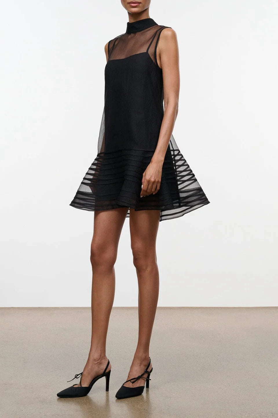 Model on a neutral background wearing black heels and the STAUD sterling mini dress in black which features a sheer outer layer with a high neck and a mini black slip underneath.