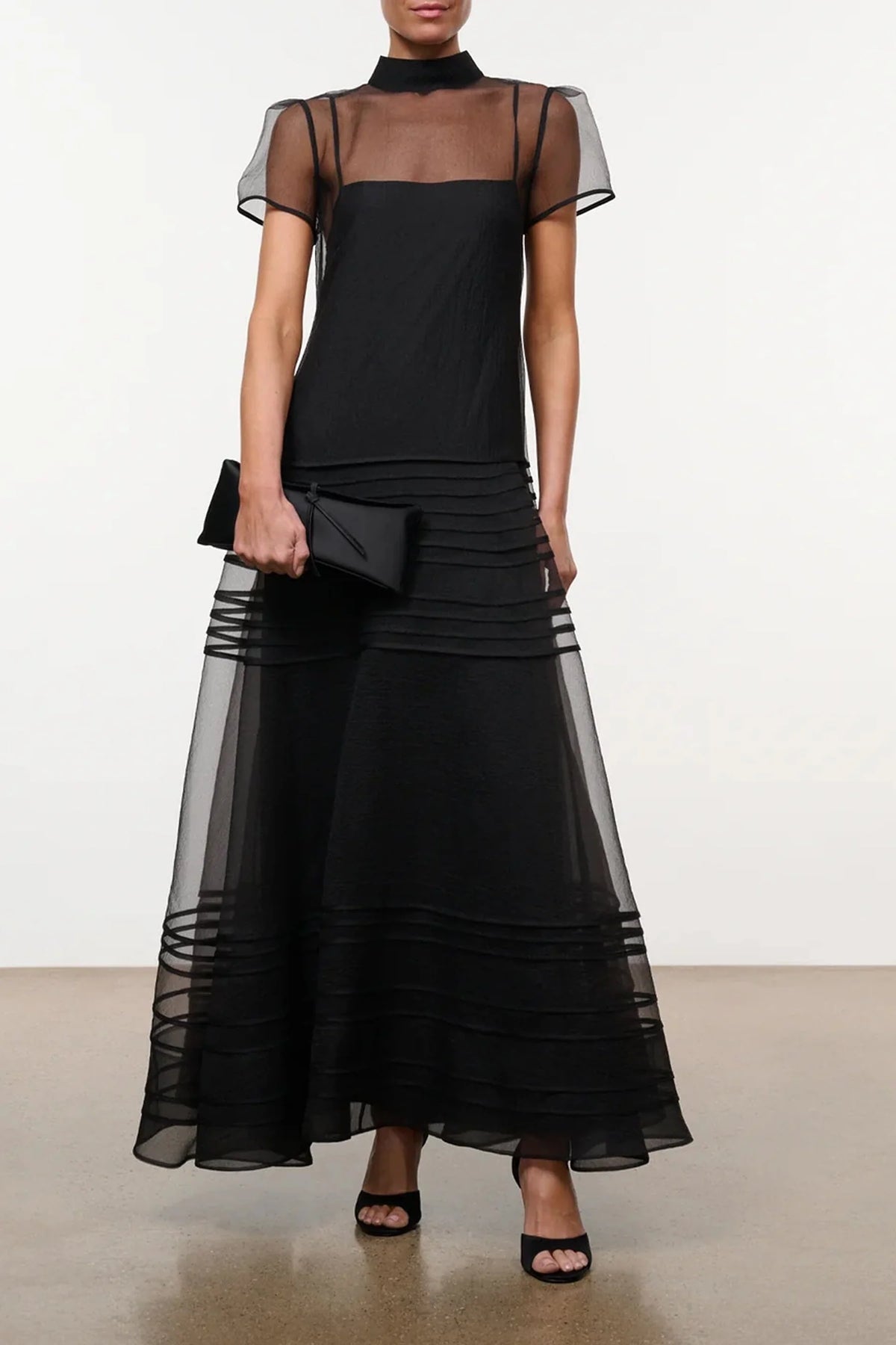 Model wearing the STAUD Stratton maxi dress that features a sheer outer layer with a black maxi slip underneath. The outer layer features a mock neckline and capped sleeves.