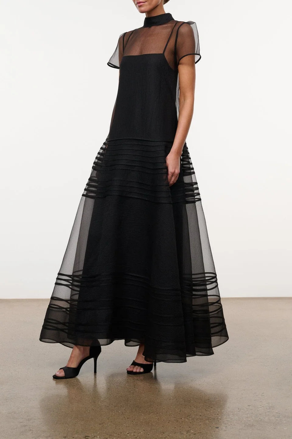 Model wearing the STAUD Stratton maxi dress that features a sheer outer layer with a black maxi slip underneath. The outer layer features a mock neckline and capped sleeves.