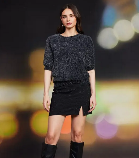 Woman wearing a grey blue textured top and skirt with black knee-high boots against a blurred colorful background
