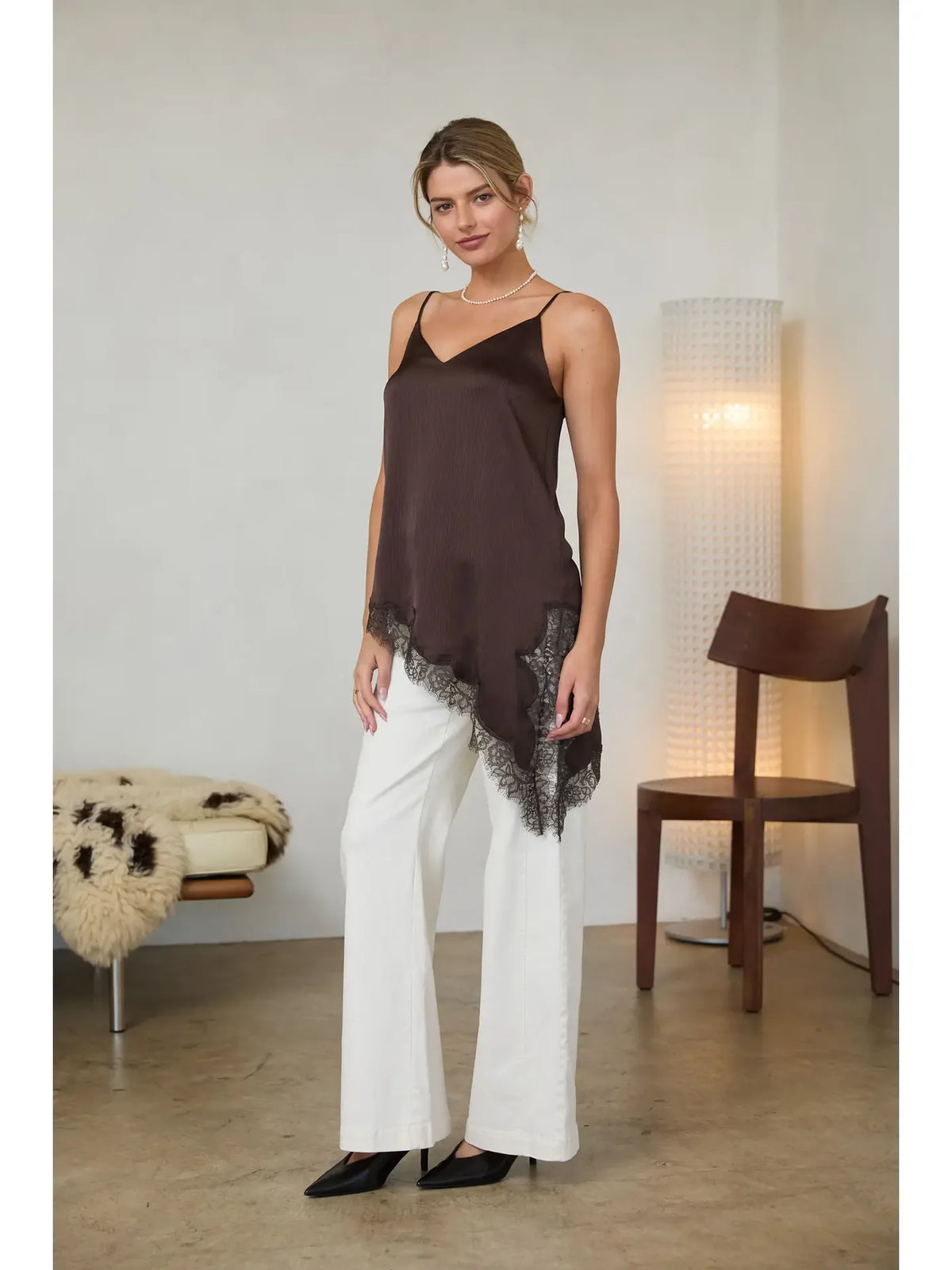 A woman models a dark brown camisole top with brown lace hem, paired with white wide-leg pants and black heels, standing in a minimalist room with a floor lamp and wooden chair.