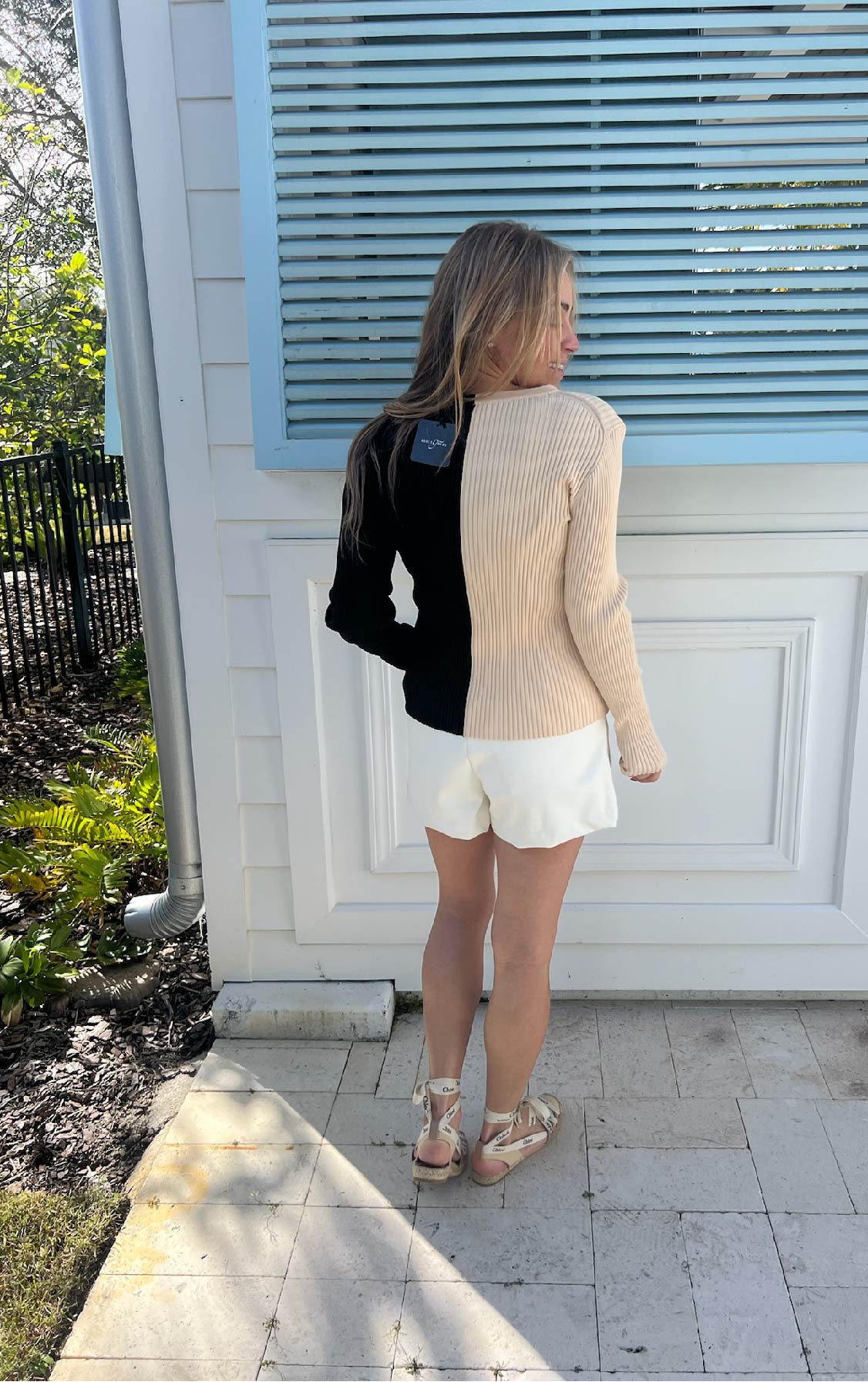 Woman wearing white shorts and a black and tan sweater standing infront of a white wall