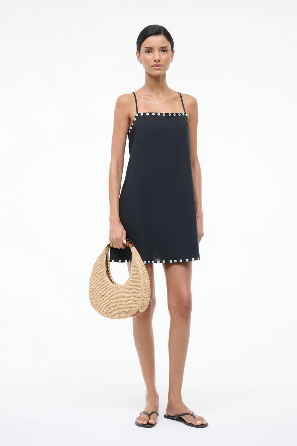 Woman wearing a black dress with white button details, holding a woven bag on a white background