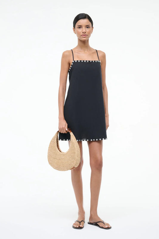 Woman wearing a black dress with white button details, holding a woven bag on a white background