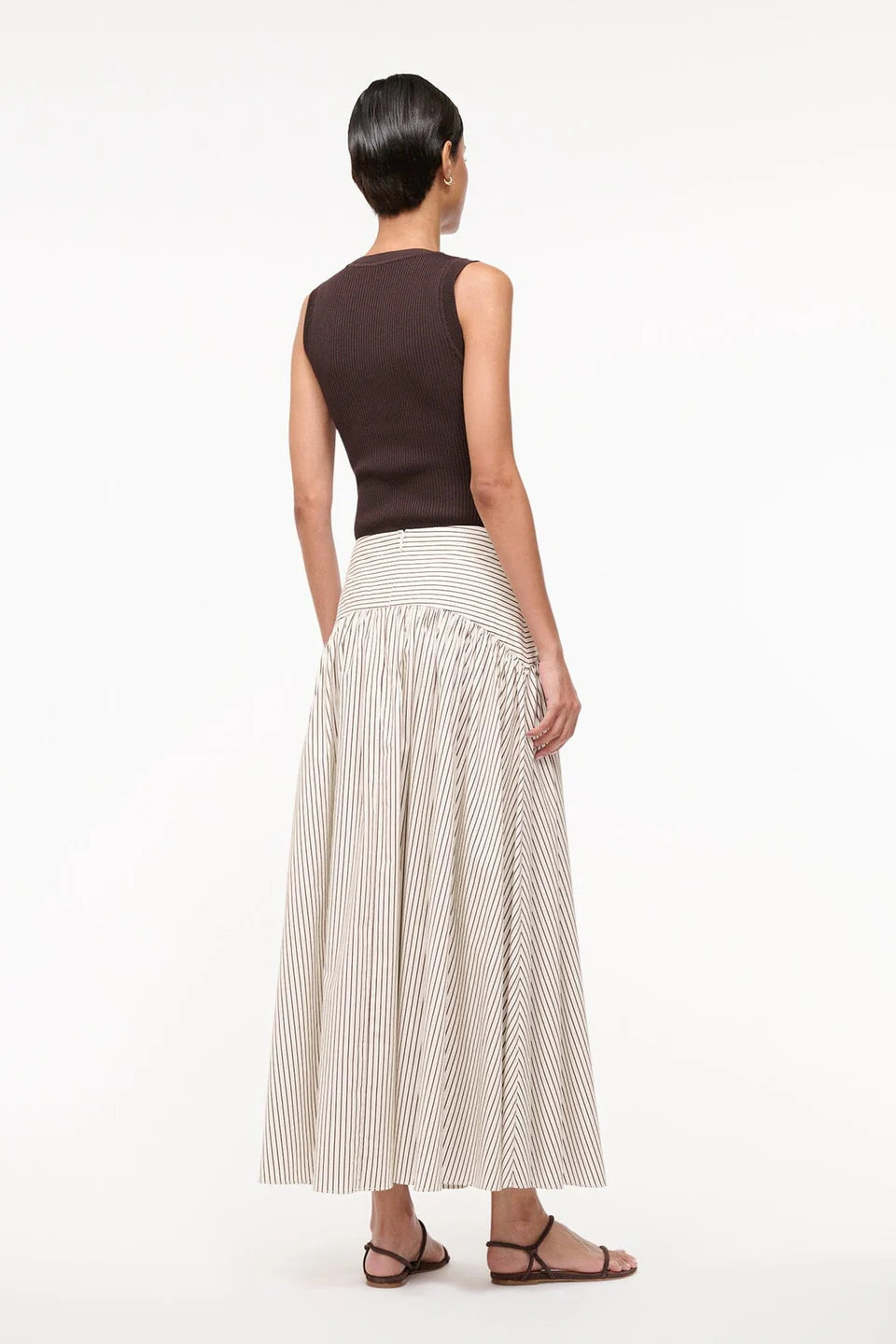 Woman wearing a sleeveless brown top and a long, light-colored pleated skirt on a white background