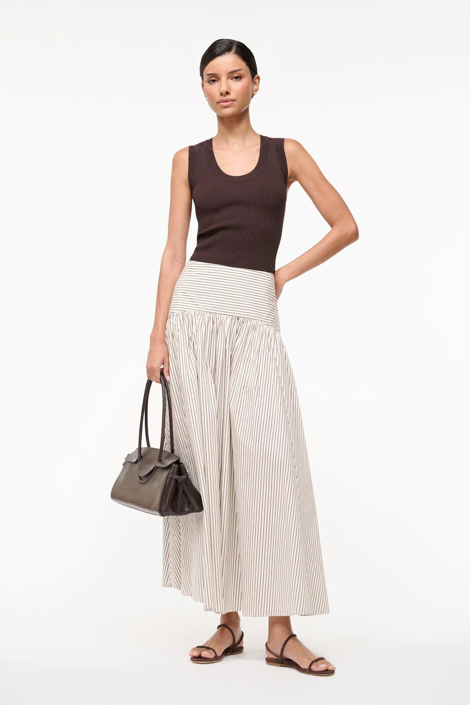 Woman wearing a brown top and white striped skirt holding a black handbag on a white background