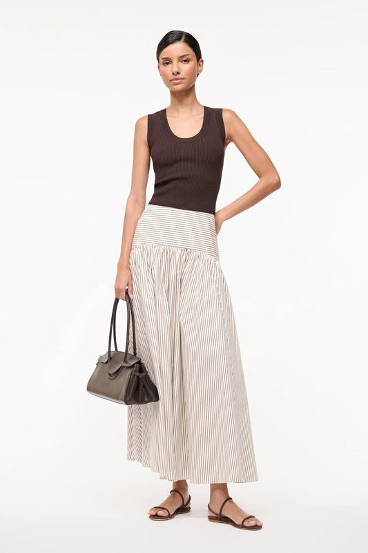 Woman wearing a brown top and white striped skirt holding a black handbag on a white background