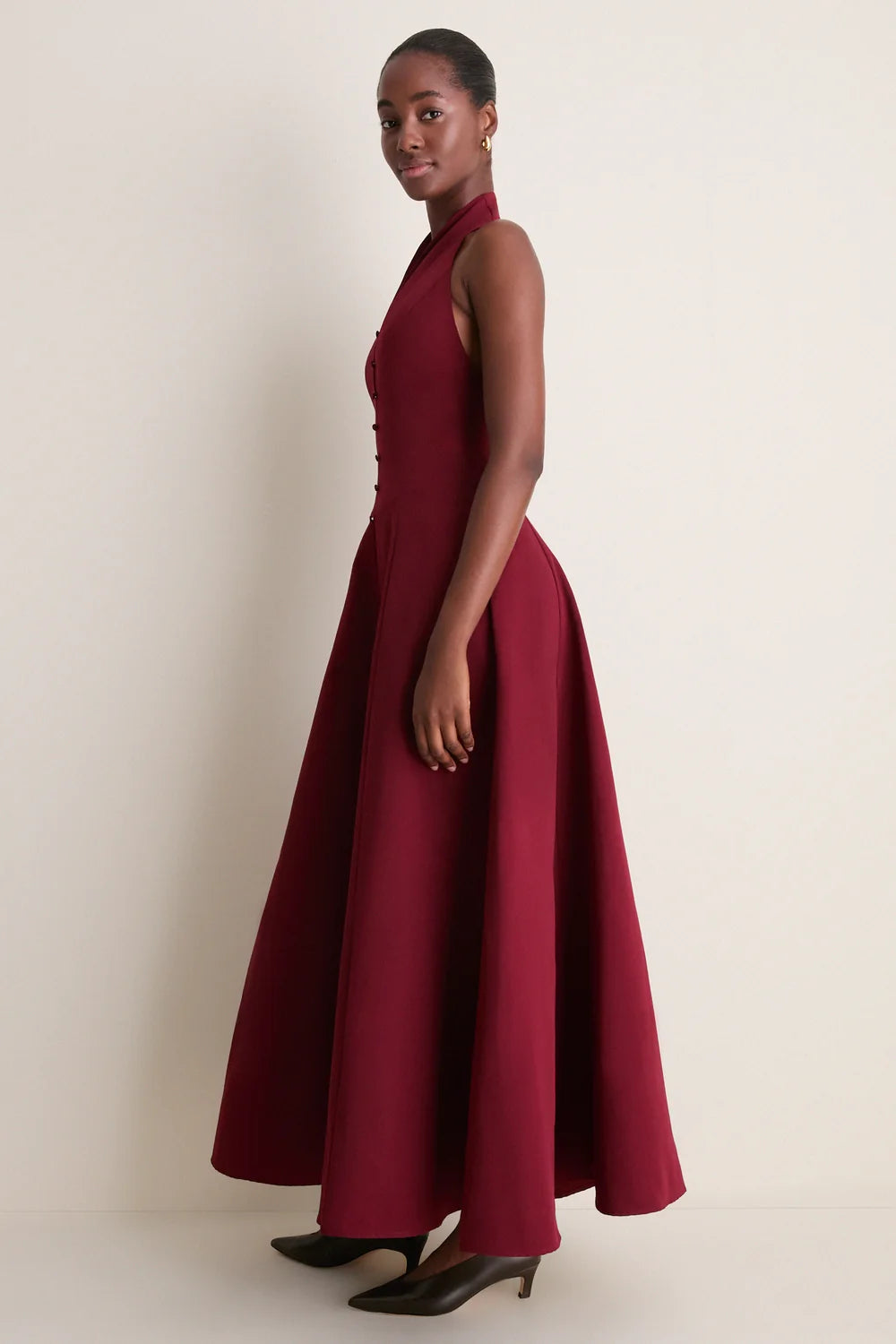 A model wearing the Staud Carmino Stella dress which features button detailing along the front wearing brown heels on a neutral background.