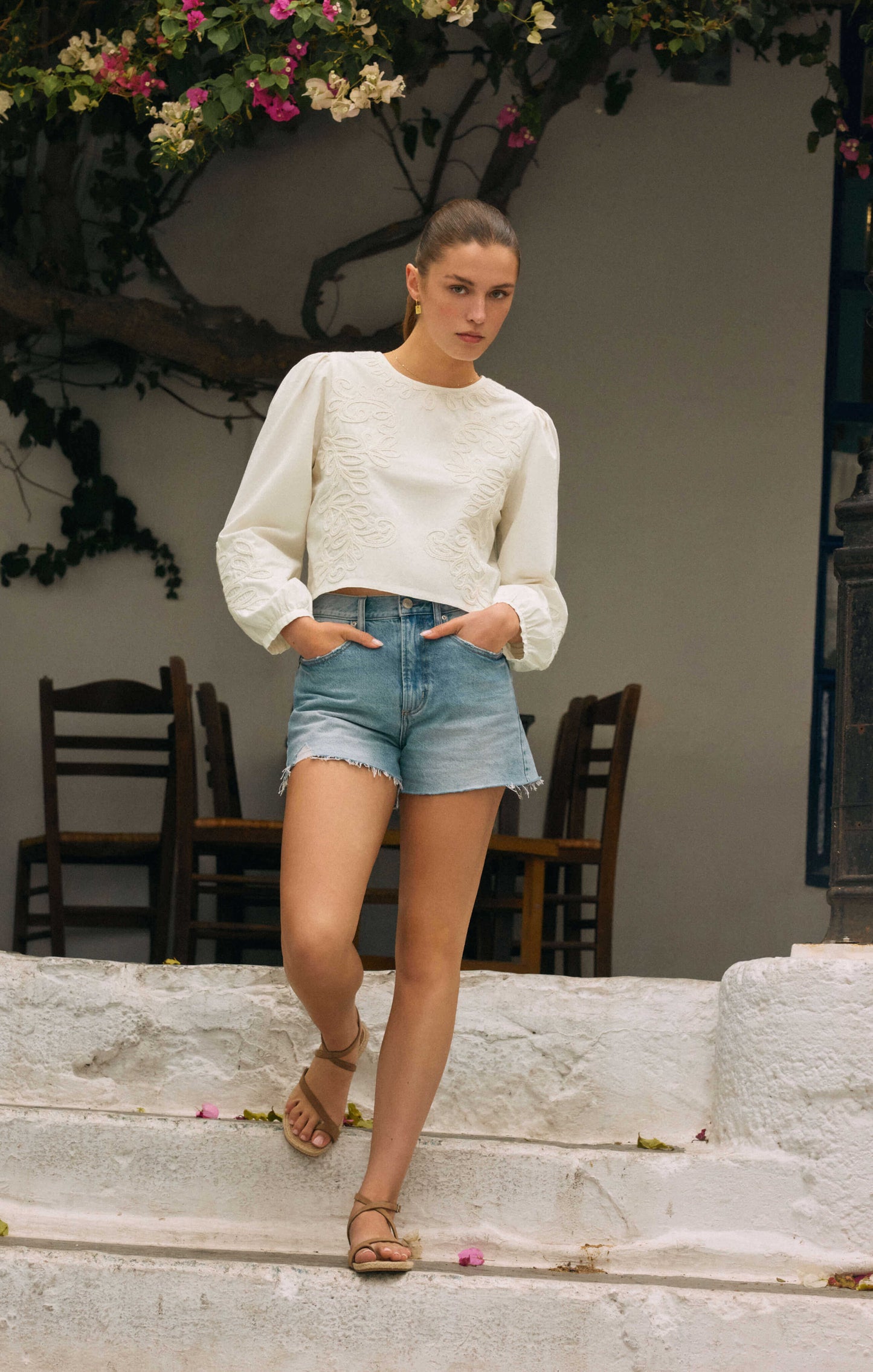 Woman in a white sweater and denim shorts standing on stone steps with greenery in the background