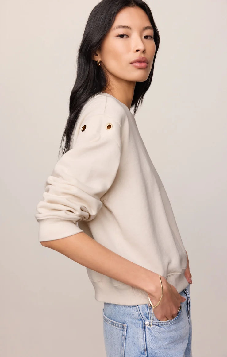 A woman wearing a cream-colored crewneck sweatshirt with decorative grommet details on the sleeve, paired with light blue jeans, photographed against a neutral light beige background.