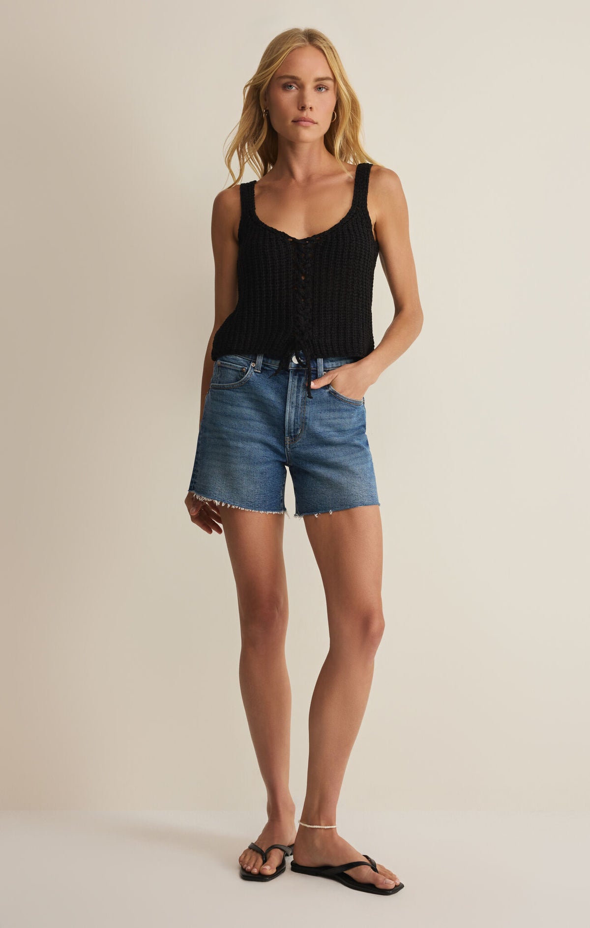 Woman wearing a black crochet tank top and blue denim shorts on a beige background