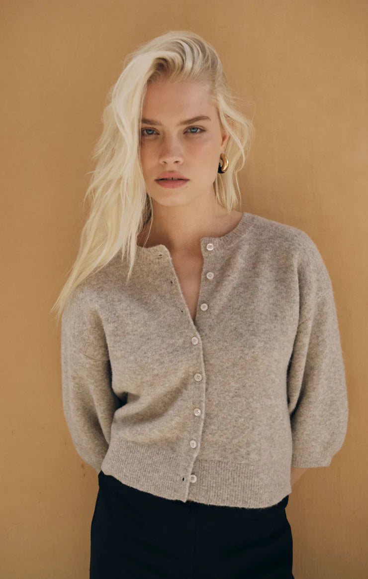 A blonde model wearing a light beige knit cardigan with a buttoned neckline, styled with black pants against a warm tan background.